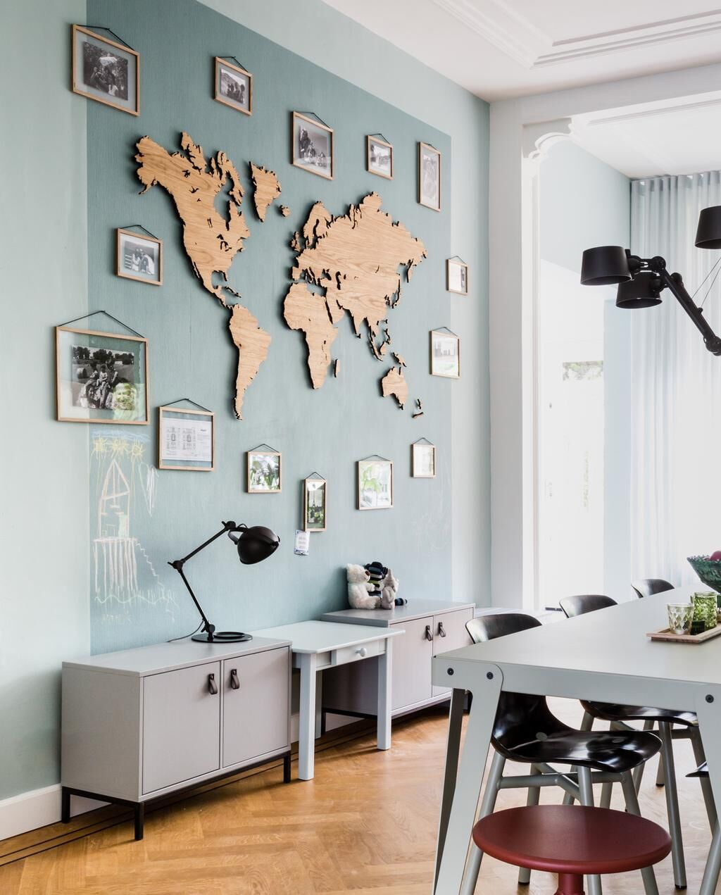 Innenansicht eines modernen Raumes mit einer Weltkarte aus Eichenholz an der Wand und mehreren eingerahmten Fotos drumherum. Eine Tischlampe steht auf einem Schrank und ein Tisch mit Stühlen befindet sich im Vordergrund.